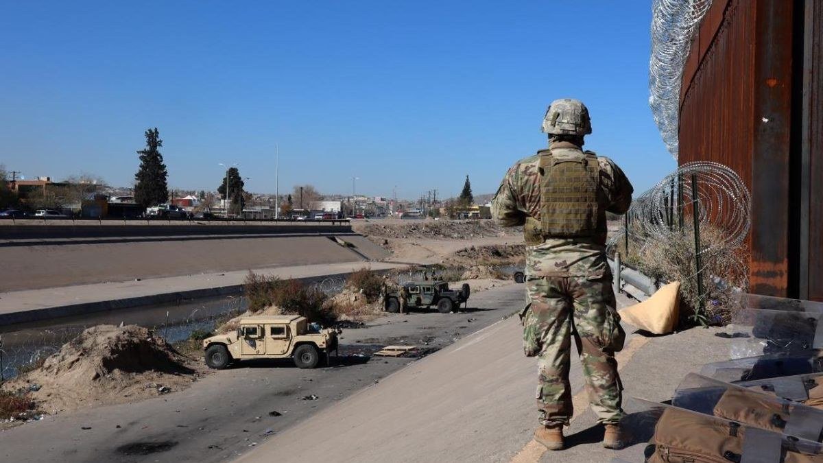 U.S. soldier in camouflage and body armor stands beside the rust-colored border wall and coiled razor wire as two Humvees sit along a concrete river channel