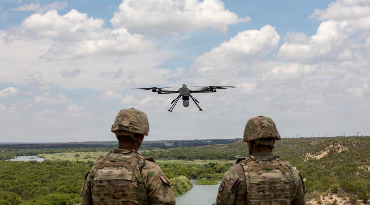 Two U.S. soldiers seen from behind watch a small quadcopter drone fly over a river valley